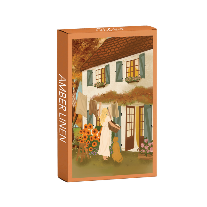 The Amber Linen | 99 Piece Jigsaw Puzzle by Olleo Puzzles features Nicolet Boon's artwork of a woman with a dog hanging laundry outside a flower-adorned cottage with green shutters.