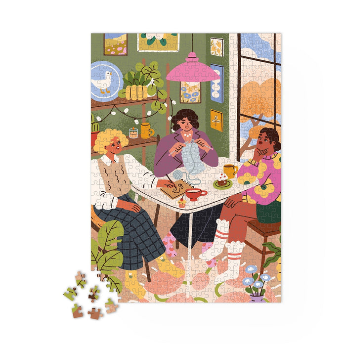Three women chat and sip tea in a cozy room as they work on the Maison Joliette "Girlfriends" 1,000-piece jigsaw puzzle. Surrounded by knitting supplies and plants, they enjoy this beautifully crafted puzzle, made in France.