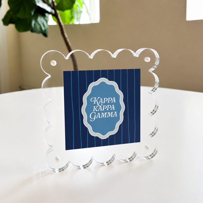 The Kappa Kappa Gamma Square Scalloped Acrylic Frame by The Darling Effect features a blue striped center with the sorority name in white, shown standing on a white table near a green plant and window.