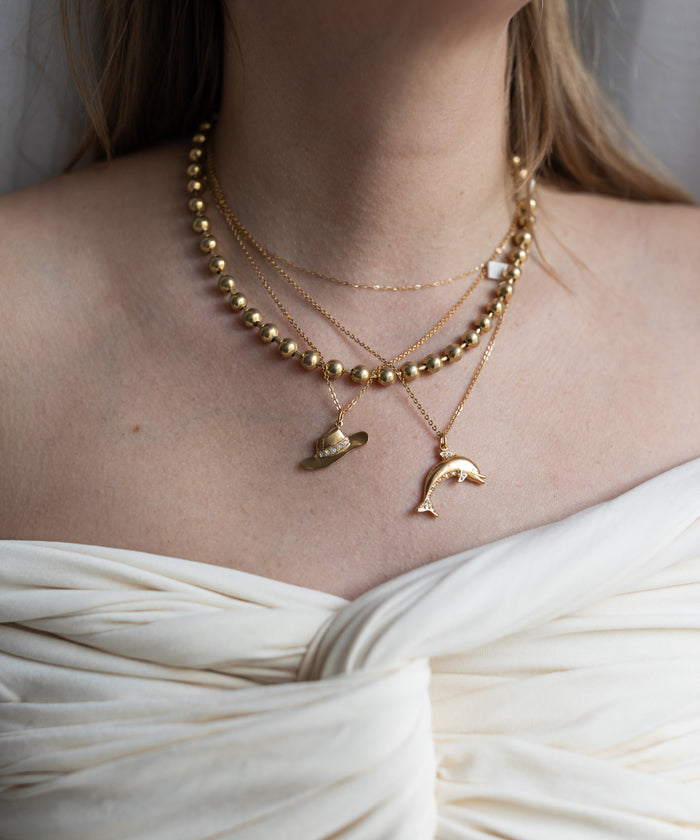 A woman wears WALD Berlin’s SAMPLE SALE Pop Cowgirl Necklace—an 18k gold plated, 55cm layered piece with hat and dolphin pendants—styled with an off-shoulder white top. Made in Germany for an elegant touch.
