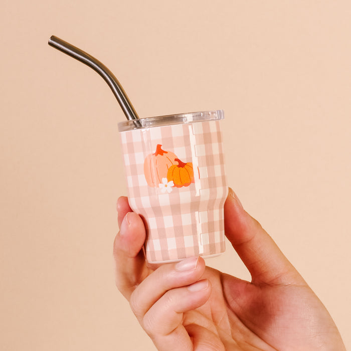 A hand holds The Darling Effect's Pumpkin Gingham 2 oz Tiny Tumbler, featuring pink and white gingham, orange pumpkin and white flower prints, set against a solid light beige background.