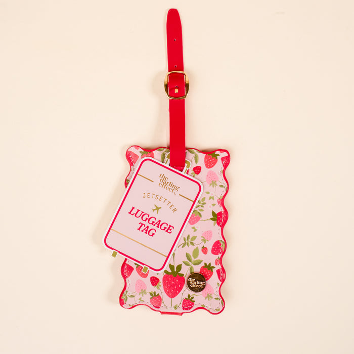 The Darling Effect's Strawberry Fields Jetsetter Luggage Tag features a pink and red strawberry pattern, a red strap with gold buckle, and a privacy flap for security. This stylish travel accessory is displayed on a light background.