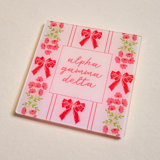 The Darling Effect Alpha Gamma Delta Acrylic Coaster features red bows and pink flowers, “alpha gamma delta” in cursive at the center, and a light background—ideal as a thoughtful sorority gift.