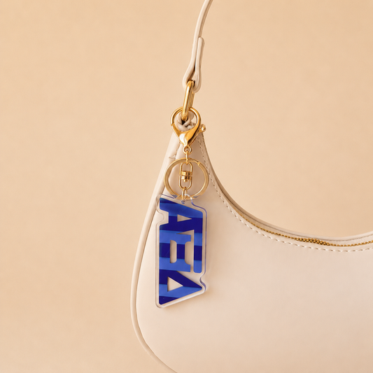 A beige handbag with gold hardware displays The Darling Effect’s Alpha Xi Delta Stripes Acrylic Keychain in blue and clear on the strap, featuring stylized Greek letters. The beige background makes it a perfect stylish sorority gift.