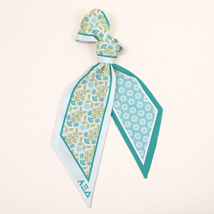 The Darling Effect’s Alpha Xi Delta Twirl & Tie Scarf Accessory showcases light blue, green, and teal floral-geometric designs with AΞΔ lettering, tied loosely for versatile styling and a sorority-inspired touch.