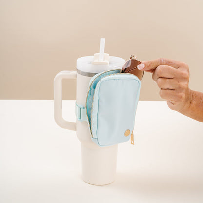 A hand places a key fob into The Darling Effect Aqua Tumbler Fanny Pack in light blue, attached to the handle of a large white tumbler with a straw, set against a neutral background.
