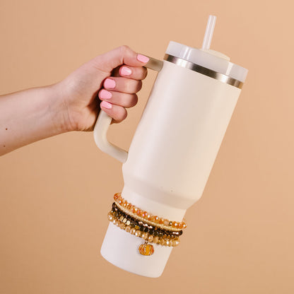 A hand with pink nail polish holds a cream 40 oz tumbler with a straw. The Darling Effect Autumn Glow Tumbler Bracelet Stack adds flair around the handle base, set against a solid beige background.