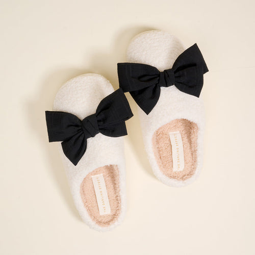 The Darling Effect's Betty Bow Black Fuzzy Slippers feature plush white material and black bows, displayed against a cream background.