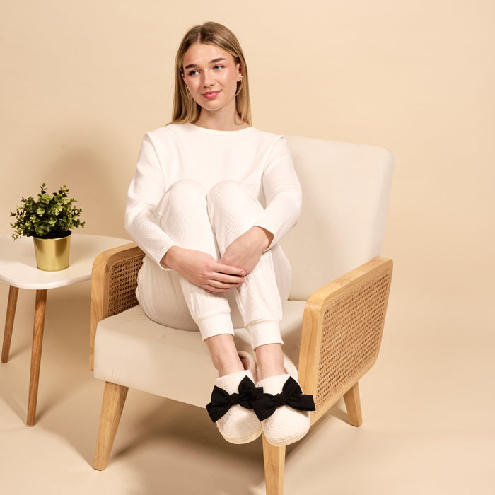 A woman in white loungewear sits on a beige armchair, wearing The Darling Effect’s Betty Bow Black Fuzzy Slippers. Her knees are up, plush slippers with black bows visible. A small table with a potted plant sits beside her against a beige background.