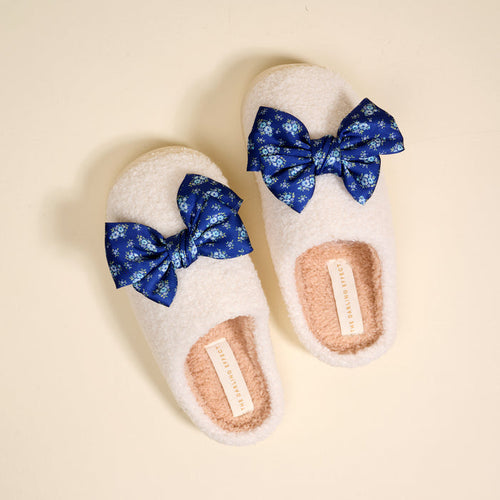 The Darling Effect Betty Bow Endless Daydream Navy Fuzzy Slippers feature white, plush comfort, blue floral bows, a tan interior, and a branded tag—making these cozy slippers both charming and comfortable.