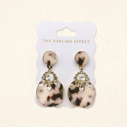 The Darling Effect’s Blonde Tortoise Show Stopper Earrings feature lightweight acetate with circular tops, large round bottoms, gold accents, and clear marquise rhinestones. Presented on a white card branded THE DARLING EFFECT.