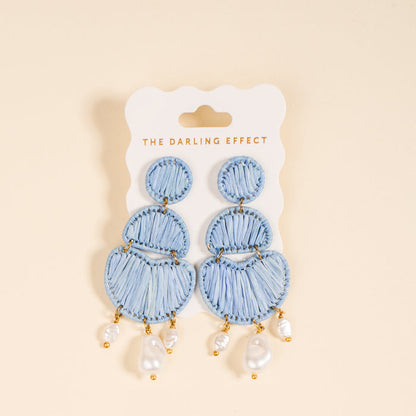 The Darling Effect’s Blue Pearl Affair Earrings are light blue, woven, tiered boho earrings with dangling pearls and gold accents—perfect for everyday wear, displayed on a card labeled The Darling Effect.