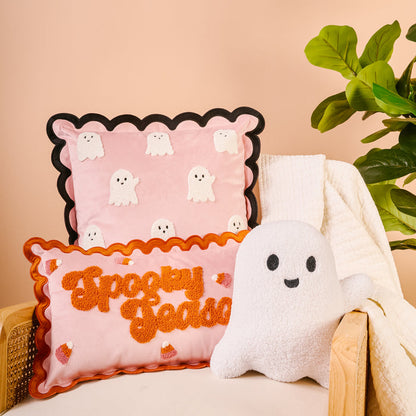 A cozy chair with a white blanket holds The Darling Effect Boo Crew Scalloped Square Throw Pillow—pink with small ghosts—plus an orange “Spooky Szn” pillow and plush ghost, next to a leafy plant against a beige background.