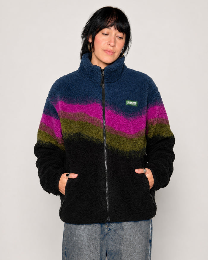 A woman wearing a fuzzy quarter-zip fleece jacket with horizontal wavy stripes in navy blue, magenta, olive green, and black, paired with light-wash jeans against a plain light gray background.

Shop Borealis High Pile Fleece Jacket Inspired by Alaskan Parks | multi-color