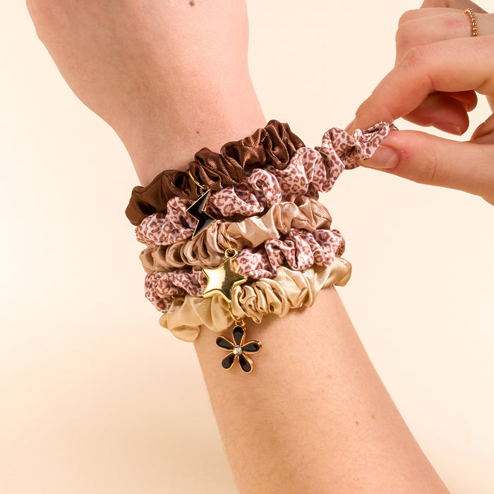 A hand and wrist showcase The Darling Effect's Born Wild Scrunchie Charm Set in brown, beige, and leopard print, each with chic star and flower charms, while another hand adjusts the top scrunchie.