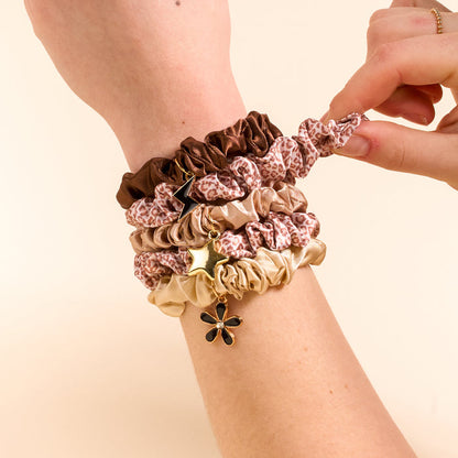 A hand and wrist showcase The Darling Effect's Born Wild Scrunchie Charm Set in brown, beige, and leopard print, each with chic star and flower charms, while another hand adjusts the top scrunchie.