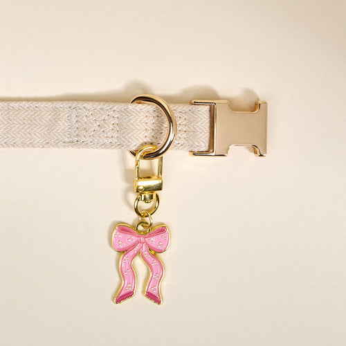 The Darling Effect's Bow Pink Pawfect Collar Charm—a pink bow-shaped charm with gold accents—adorns a beige fabric collar with a gold buckle and D-ring, adding stylish flair to your pet accessory collection.