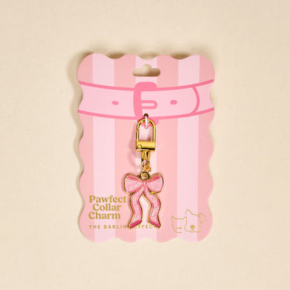 The Darling Effect's Bow Pink Pawfect Collar Charm features a pink bow shape with gold accents and a gold clasp, displayed on pink and white striped packaging—an adorable accessory for any pet collar charm collection.
