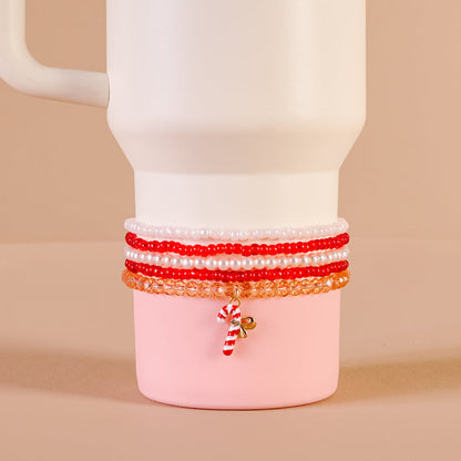 The Darling Effect's Candy Cane Crush Tumbler Bracelet Stack in red, white, and orange with a candy cane charm decorates a white and pink mug. Unique tumbler accessory shown on a light beige background.