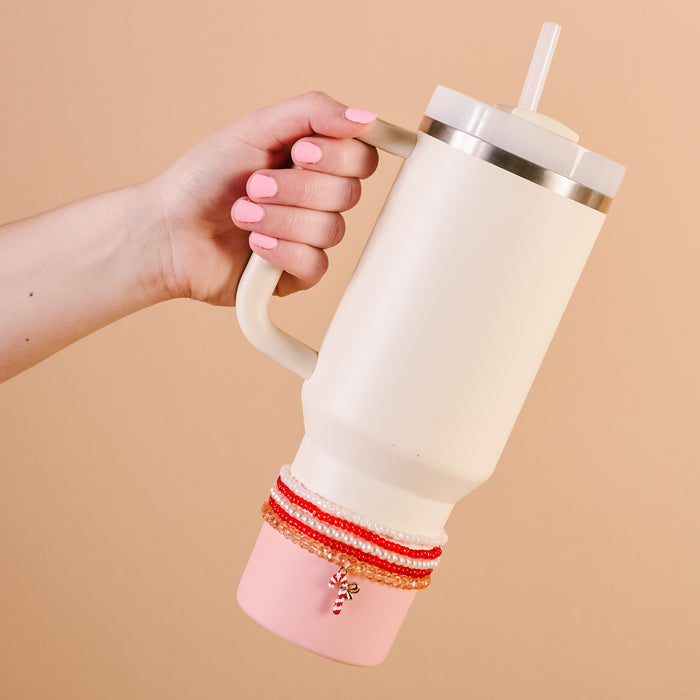 A hand with pink nails holds The Darling Effect’s Candy Cane Crush Tumbler Bracelet Stack adorning a large pink 40 oz tumbler with a straw. A candy cane charm hangs from the stack against a simple beige background.