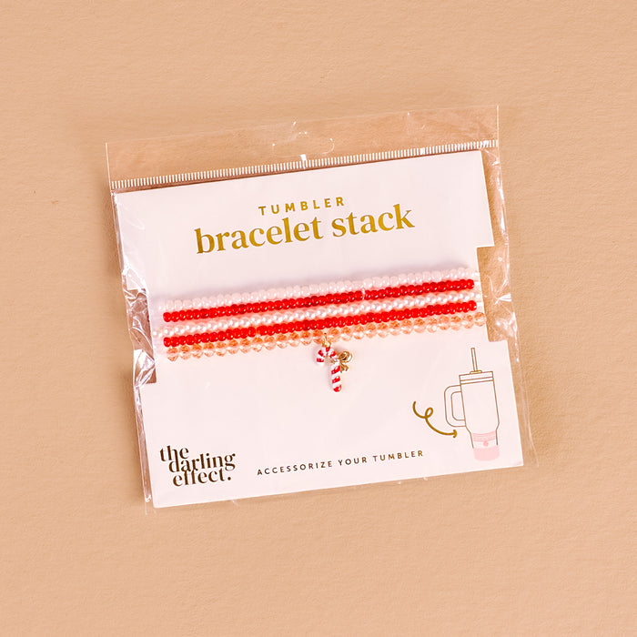 The Darling Effect's Candy Cane Crush Tumbler Bracelet Stack features red, pink, and white beaded bracelets with a candy cane charm, packaged with a tumbler illustration—ideal for tumbler accessory fans.