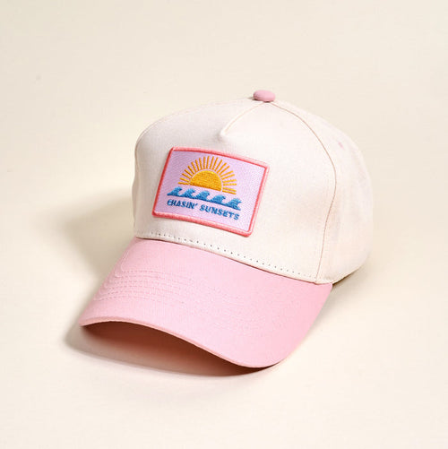 The Darling Effect’s Chasin’ Sunsets Classic Canvas Hat features a beige canvas design with a pink brim and coordinating top button, making it ideal for everyday wear. The relaxed-fit baseball cap is adorned with a front patch depicting a sun setting over water alongside the words “CHASIN SUNSETS.”.