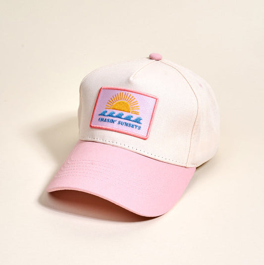 The Darling Effect’s Chasin’ Sunsets Classic Canvas Hat features a beige canvas design with a pink brim and coordinating top button, making it ideal for everyday wear. The relaxed-fit baseball cap is adorned with a front patch depicting a sun setting over water alongside the words “CHASIN SUNSETS.”.