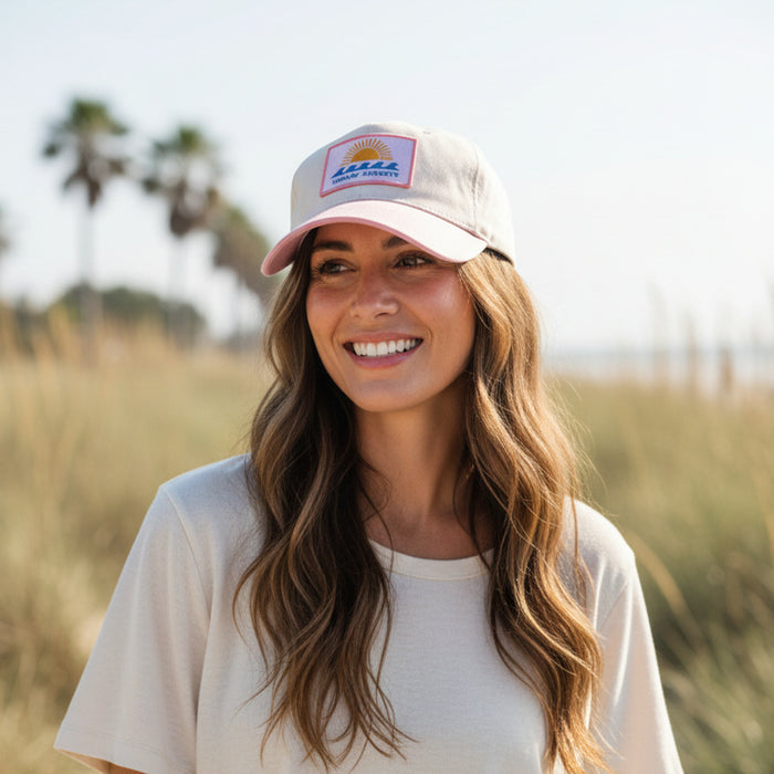 A woman with long wavy hair smiles while standing outdoors in a grassy area with palm trees in the background, embracing the comfort of everyday wear in The Darling Effect’s Chasin’ Sunsets Classic Canvas Hat.