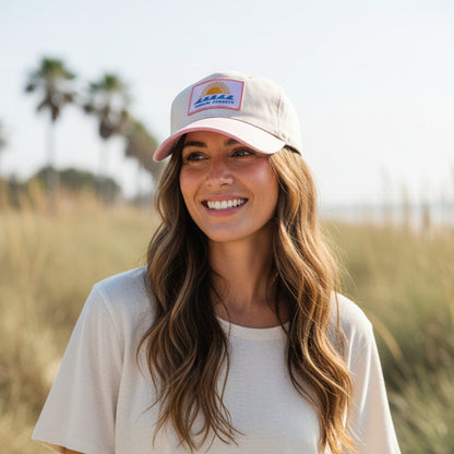 A woman with long wavy hair smiles while standing outdoors in a grassy area with palm trees in the background, embracing the comfort of everyday wear in The Darling Effect’s Chasin’ Sunsets Classic Canvas Hat.