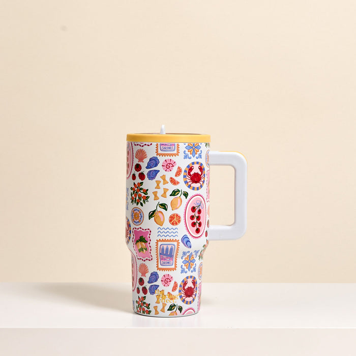 The Darling Effect's Ciao Bella 20 oz Lifestyle Flip Straw Tumbler, featuring a large handle and colorful fruit and floral patterns, sits on a white surface with a beige background.