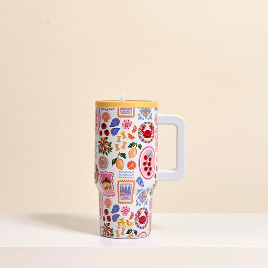 The Darling Effect's Ciao Bella 20 oz Lifestyle Flip Straw Tumbler, featuring a large handle and colorful fruit and floral patterns, sits on a white surface with a beige background.