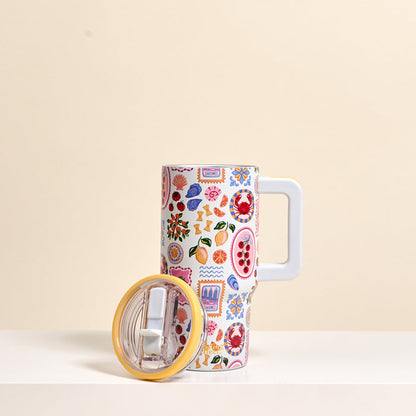 The Darling Effect Ciao Bella 20 oz Lifestyle Flip Straw Tumbler features a vibrant fruit and geometric pattern. It stands upright on a white surface, with its clear lid featuring a yellow rim beside it, set against a light beige background.