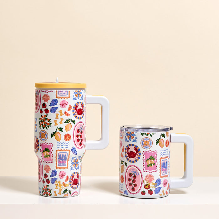 Two white mugs with playful fruit and flower designs sit side by side. The taller one is The Darling Effect's Ciao Bella 20 oz Lifestyle Flip Straw Tumbler with a yellow lid; the shorter mug is open on a beige background.