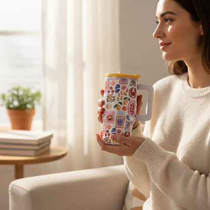 A woman in a cream sweater sits on a couch holding The Darling Effect's Ciao Bella 20 oz Lifestyle Flip Straw Tumbler featuring vibrant patterns. A small potted plant and stacked books are on a table in the softly lit background.