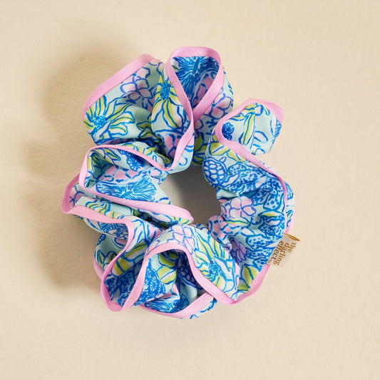 The Darling Effect's Coastal Cove Ruffle Up Scrunchie, a blue, green, and pink floral oversized scrunchie with light pink trim and a small tan tag, is shown on a beige background and is gentle on hair.