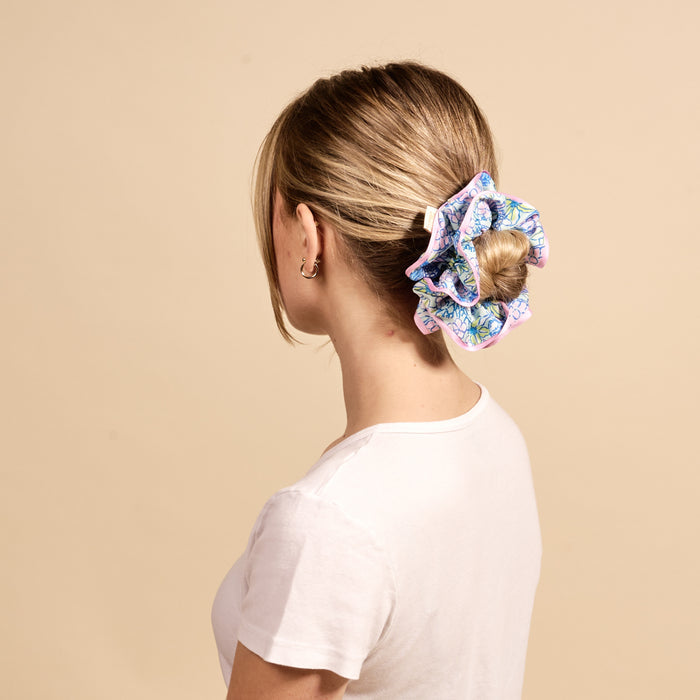 A woman with blonde hair in a low bun, secured with The Darling Effect’s Coastal Cove Ruffle Up Scrunchie, is shown from behind. She wears a plain white t-shirt and small hoop earrings against a beige background.