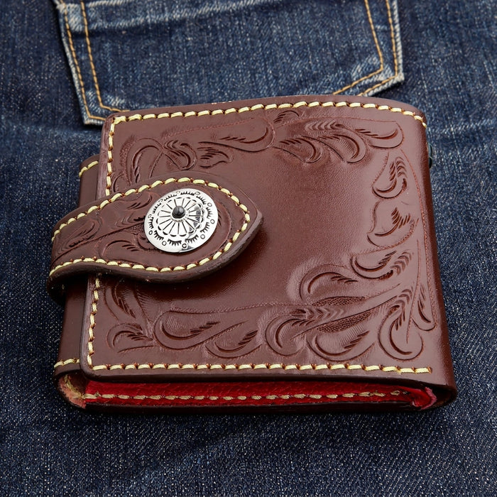 Closed front view of a dark brown hand-tooled biker wallet on blue denim, concho left of center and a red interior edge.
