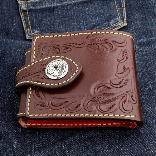 Closed front view of a dark brown hand-tooled biker wallet on blue denim, concho left of center and a red interior edge.