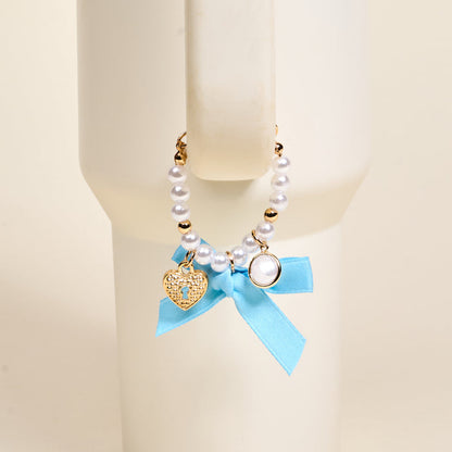 The Darling Effect Daydreamer Tumbler Charm features pearls, gold beads, a heart-shaped lock charm, and a blue ribbon bow, making it an adorable accessory for your favorite drinkware.