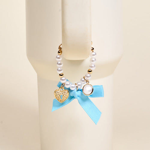 The Darling Effect Daydreamer Tumbler Charm features pearls, gold beads, a heart-shaped lock charm, and a blue ribbon bow, making it an adorable accessory for your favorite drinkware.