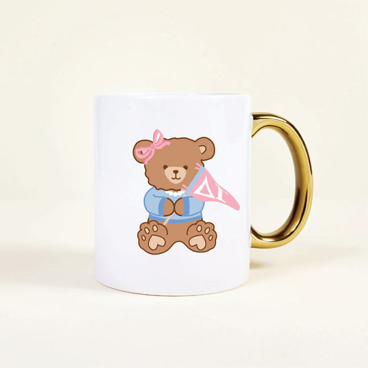 The Darling Effect's Delta Gamma 11 oz Cozy Cute Ceramic Mug features a white ceramic body, gold handle, and a sweet teddy bear in a pink bow and blue shirt holding a pink flag with the Greek letter—perfect for sorority members.