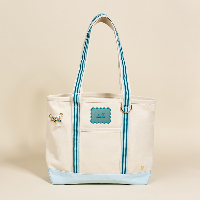 The Darling Effect Delta Zeta Canvas Tote Bag features a cream canvas, blue accents, double blue-striped handles, an AZ patch on the front, and a beaded charm—an ideal sorority bag set against a plain background.