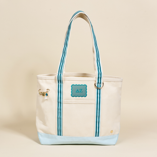 The Darling Effect Delta Zeta Canvas Tote Bag features a cream canvas, blue accents, double blue-striped handles, an AZ patch on the front, and a beaded charm—an ideal sorority bag set against a plain background.