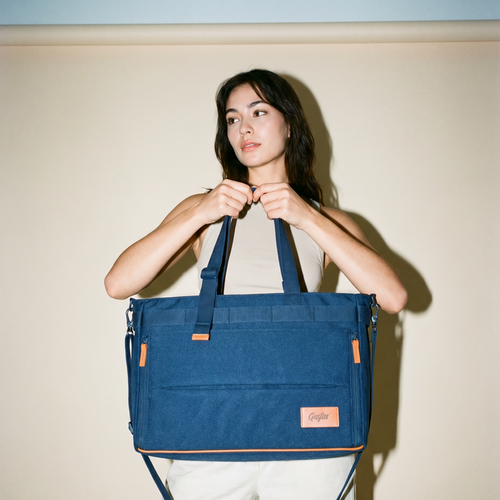 A woman holding DeskMate, a large navy blue tote bag with brown accents and a matching shoulder strap, set against a plain beige wall.