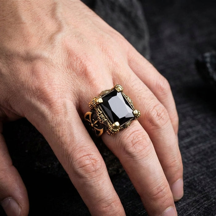 Men's dragon claw brass ring featuring a large rectangular black onyx gemstone with ornate Gothic detailing and fleur-de-lis accents, shown worn on a male hand against a dark textured background