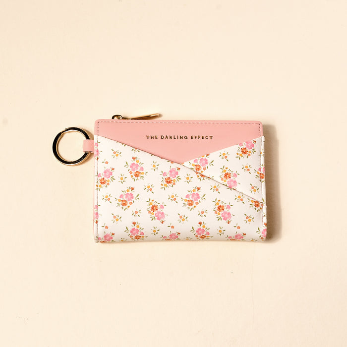 The Endless Daydream Cream Luxe Pocket Wallet by The Darling Effect is a compact vegan leather wallet with pink trim, gold zipper, keyring, and THE DARLING EFFECT logo on front. It features a pink and white envelope design with a delicate pink flower pattern.
