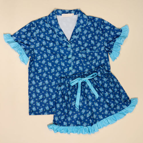 The Darling Effect’s Endless Daydream Navy Slumber Party Pajama Shorts Set features ultra-soft fabric, a floral pattern, light blue ruffled trim, a relaxed fit, short-sleeve button-up top with pocket, and matching ribbon-tie shorts.