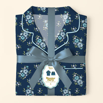 The Darling Effect’s Endless Daydream Navy Slumber Party Pajama Shorts Set features folded, ultra-soft navy pajamas with a light blue floral pattern, tied with a satin ribbon and bow. Gift-ready, it includes a tag with an illustration of the set.