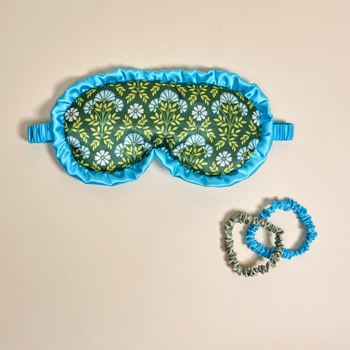 The Darling Effect's Everbloom Forest Green Beauty Sleep Mask, featuring a blue and green floral satin design with a blue ruffle, is paired with two matching satin scrunchies and elegantly displayed on a beige background.