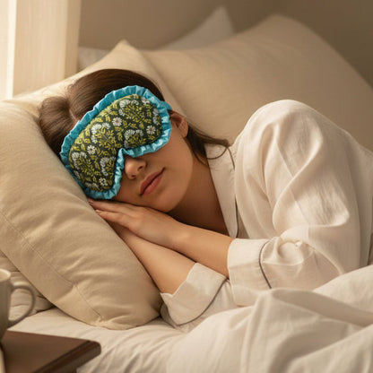 A person in a white pajama top and The Darling Effect’s Everbloom Forest Green Beauty Sleep Mask lies on their side in bed, head on a pillow with hands folded under cheek. A cup, book, and satin scrunchies rest on the bedside table.